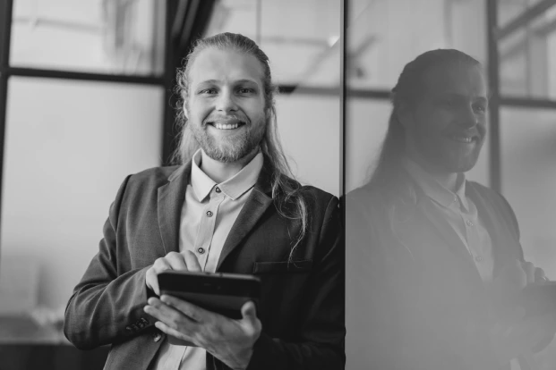 A smiling man with long hair and a beard, dressed in a suit, holding a tablet. He is standing next to a reflective glass surface, which shows his reflection. The image is in black and white.